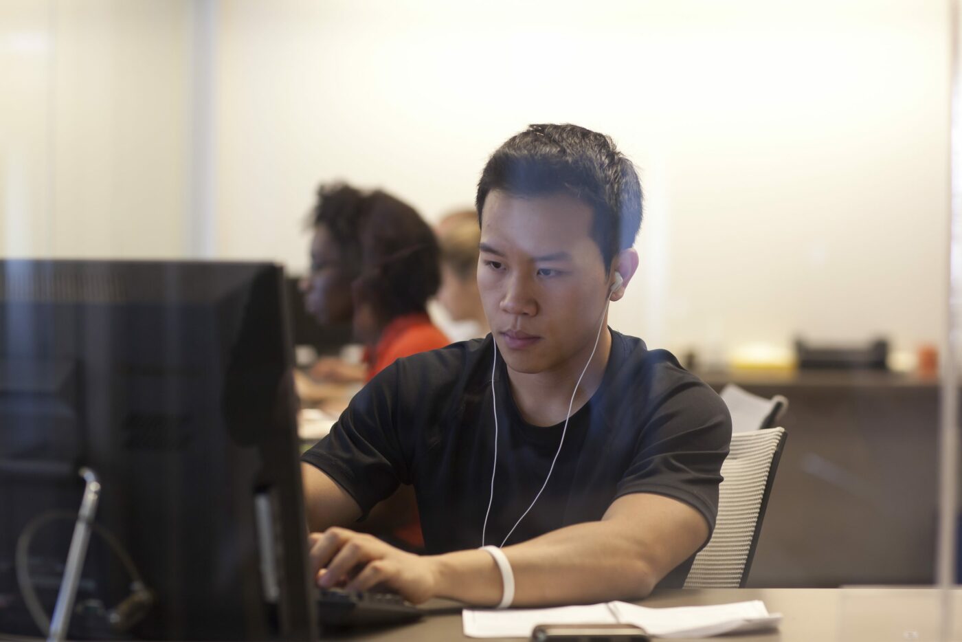 A man wearing earphones works on a computer at his desk in the Adler University Online Campus office, with other people in the background.