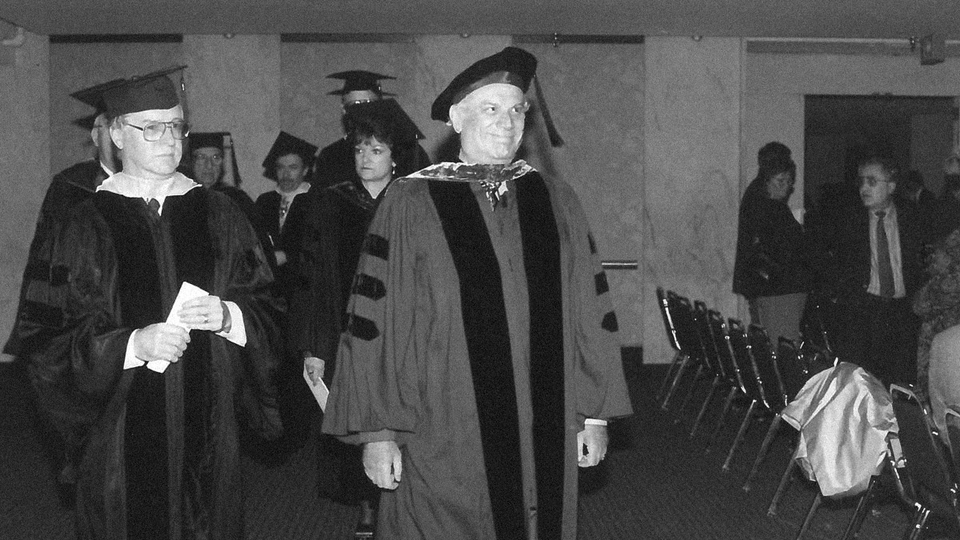 A group of people in academic regalia walk down an aisle in a formal indoor setting, likely during a graduation ceremony, honoring founders and remembering founders who shaped the institution's history.