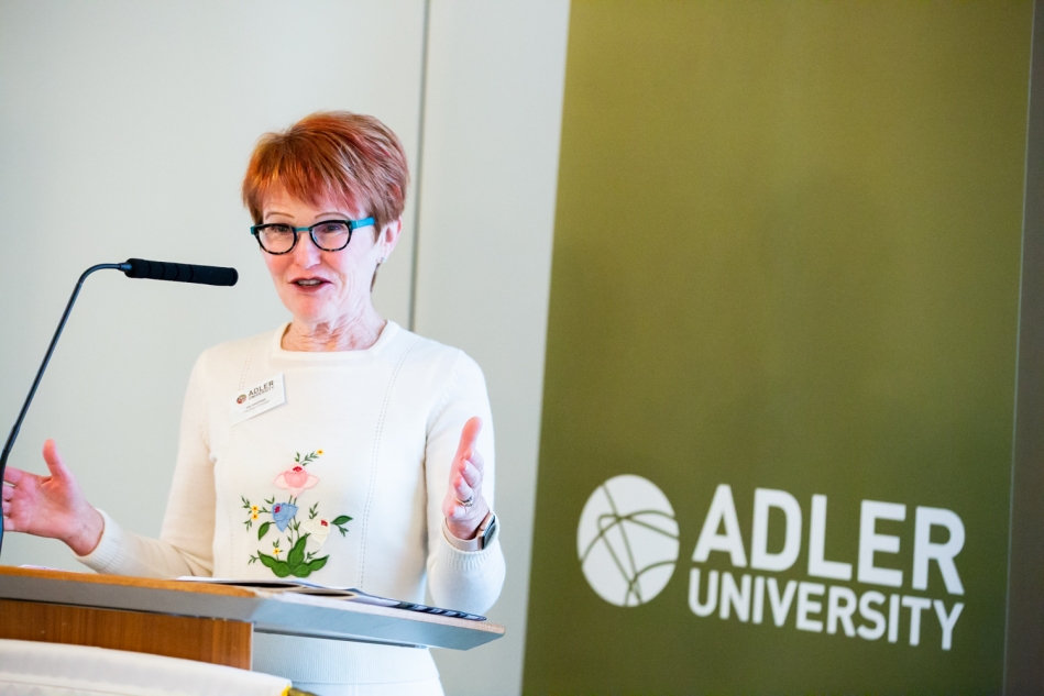 A woman speaks at a podium next to a green sign displaying the Adler University logo and name, marking a Changing of the Guard moment for the institution.