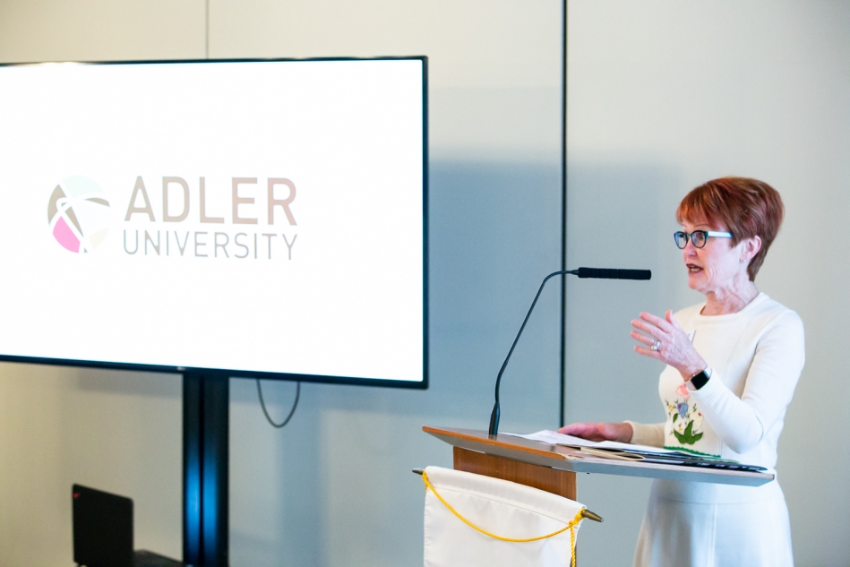 A woman speaks at a podium with a microphone during the Changing of the Guard; a screen next to her displays the Adler University logo.