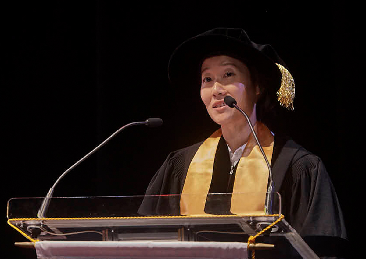 A graduate in academic regalia speaks at a podium with two microphones against a dark background at Adler University's Vancouver Campus.