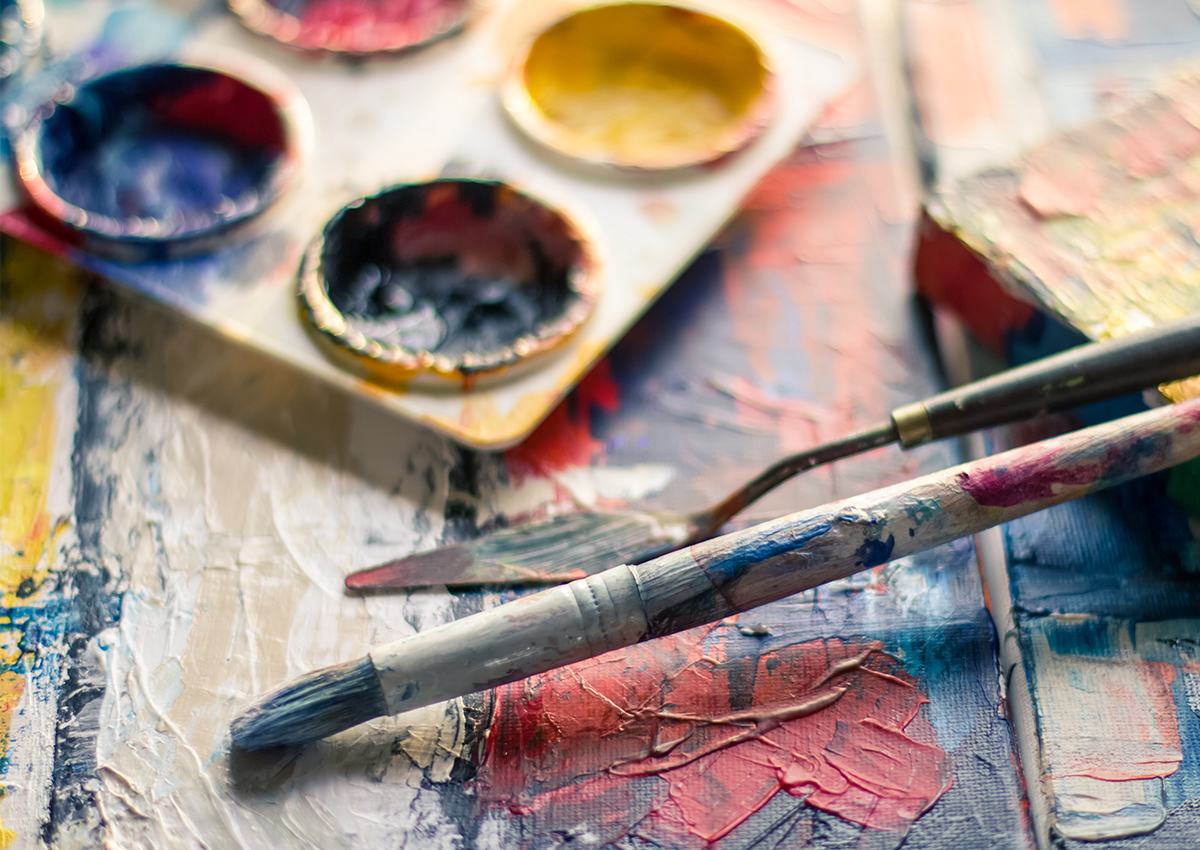 A close-up of a paintbrush and palette knife resting on a textured, colorful painting, with a paint palette in the background—reflecting creative expression often explored in an Art Therapy Program such as Adler’s renowned Ph.D. offerings.