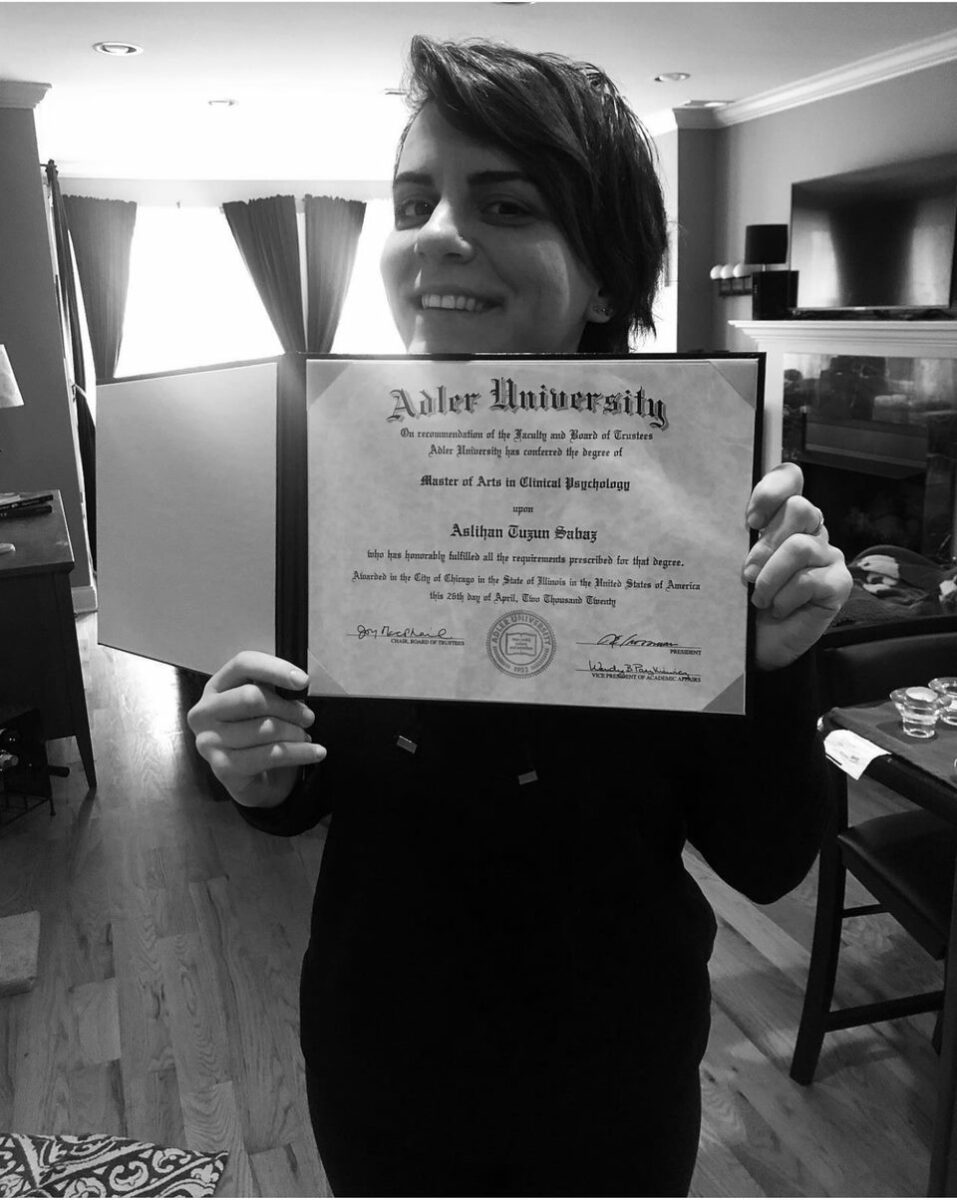 A person smiling and holding an Adler University Master of Arts in Clinical Psychology diploma, proud to support immigrant women, in a well-lit living room.