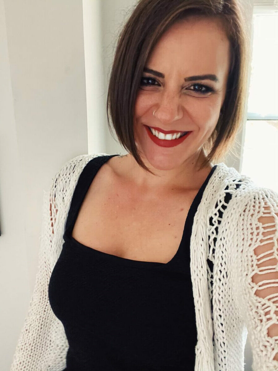 Woman with short brown hair wearing a black top and white knit cardigan, smiling at the camera in a brightly lit room in Istanbul, celebrating the strength of immigrant women.