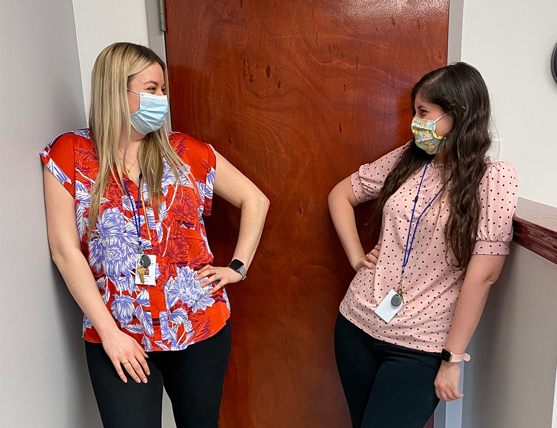 Two Adler students wearing face masks and work badges stand apart in a hallway, smiling through their eyes as they discuss how best to counsel youth during the pandemic.