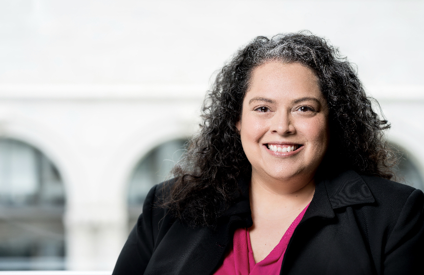 Erika Bachner sits in front of a large window with buildings blurred in the background. She is wearing a blouse and blazer.