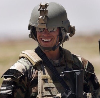 A smiling Central Illinois veteran in camouflage gear and helmet stands outdoors, wearing sunglasses and holding a rifle.