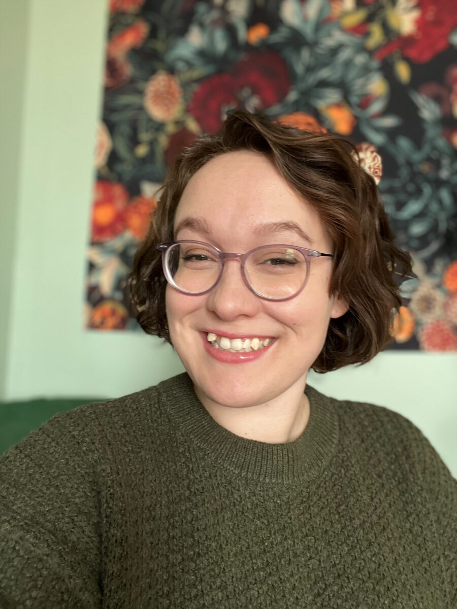 A smiling person with curly hair and glasses wears a green sweater. A colorful floral tapestry hangs in the background, creating a cheerful atmosphere.