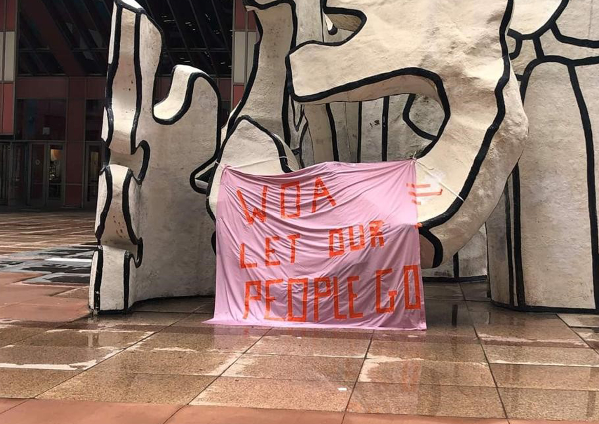 A pink banner with orange letters reading "WOA LET OUR PEOPLE GO" hangs on a large abstract black and white sculpture in an urban plaza, calling attention to prison advocacy and protection in prisons.