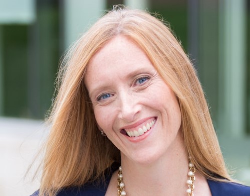 A woman with long blonde hair and a necklace smiles at the camera in an outdoor setting, celebrating as an Online Campus Graduate.