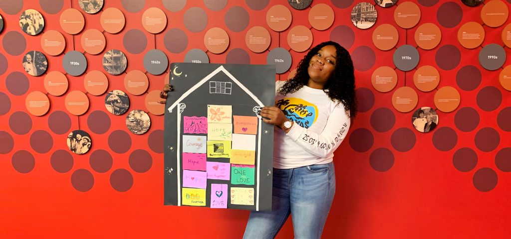 A woman stands in front of a red polka dot wall, holding a large house-shaped board decorated with colorful sticky notes and drawings, showcasing how the community unites as a student raises funds for Hurricane Dorian relief.