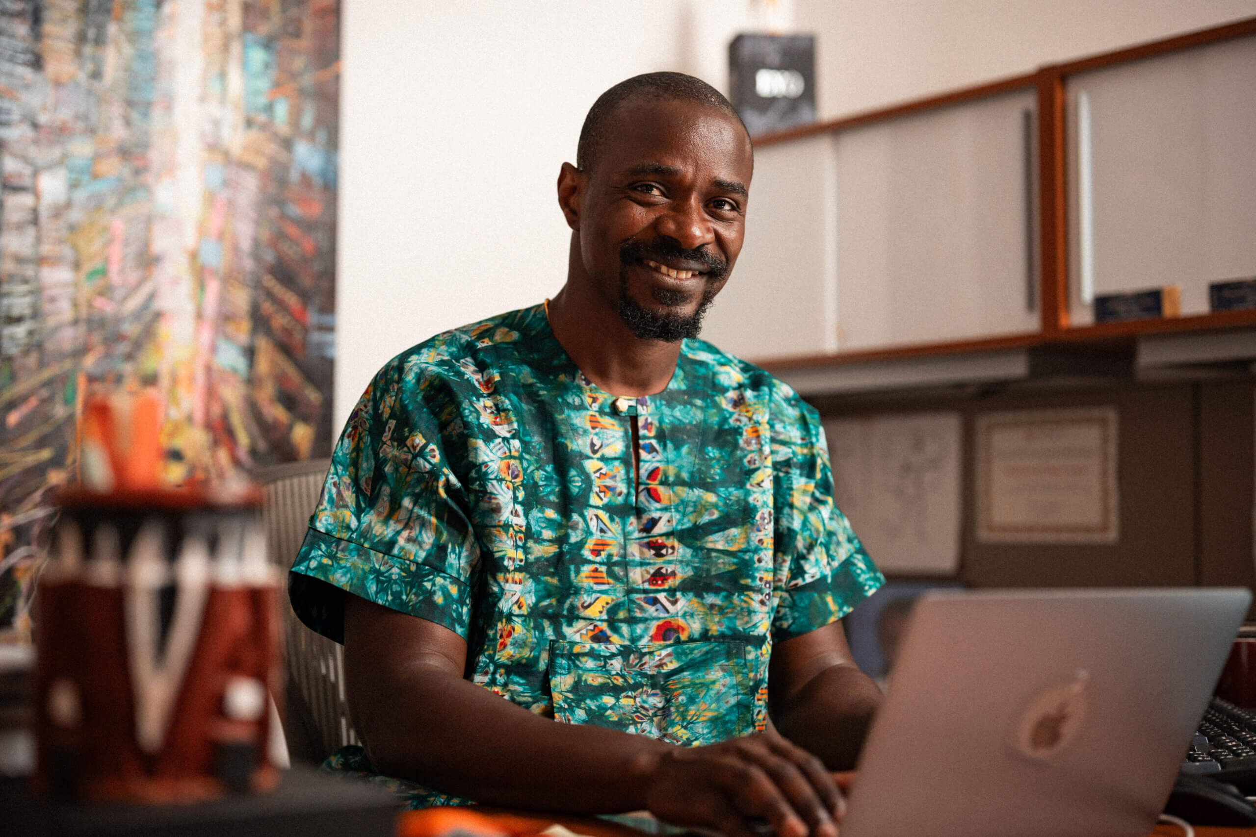 A man in a colorful patterned shirt smiles while working on a laptop at his desk in an office setting, with certificates and artwork visible in the background.