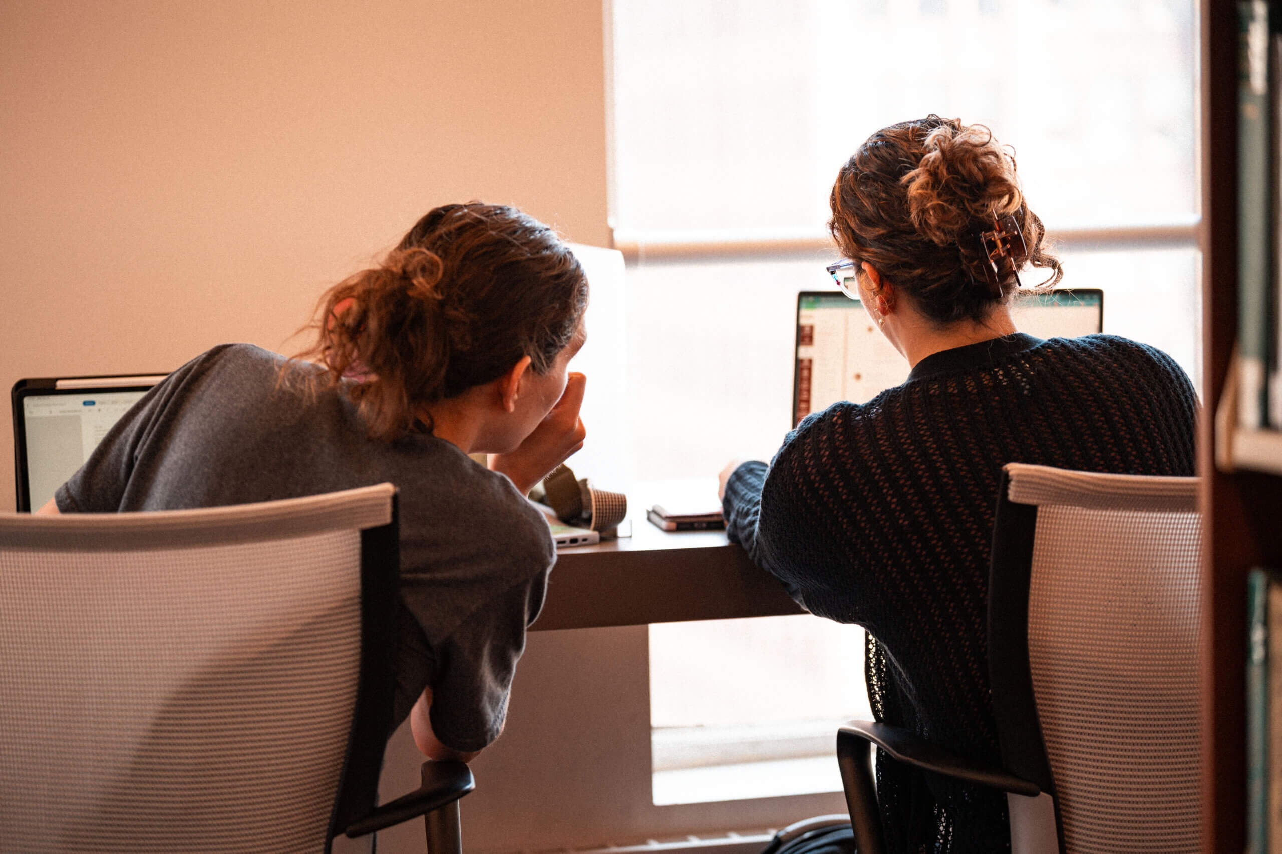 Two people with long hair in ponytails sit side by side at a desk, looking at laptop screens. One gestures toward the others screen. They are seated on white chairs in a well-lit room with a window.