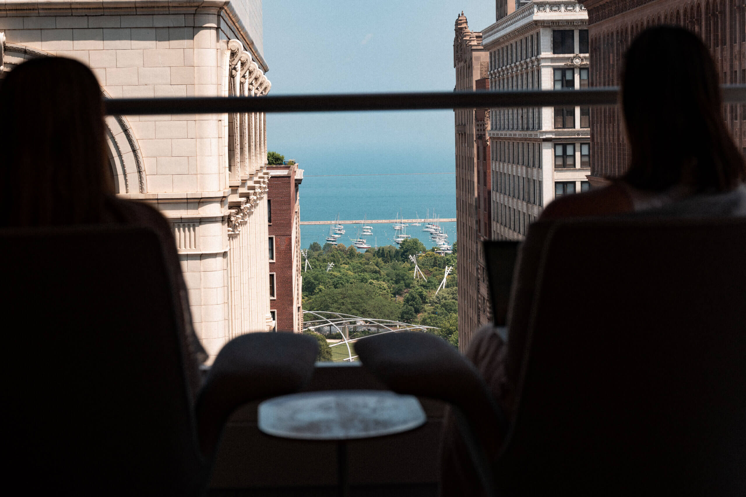 Two people sit in chairs by a window overlooking a cityscape with historic buildings, a park with trees, and a lake with sailboats in the distance under a clear blue sky.