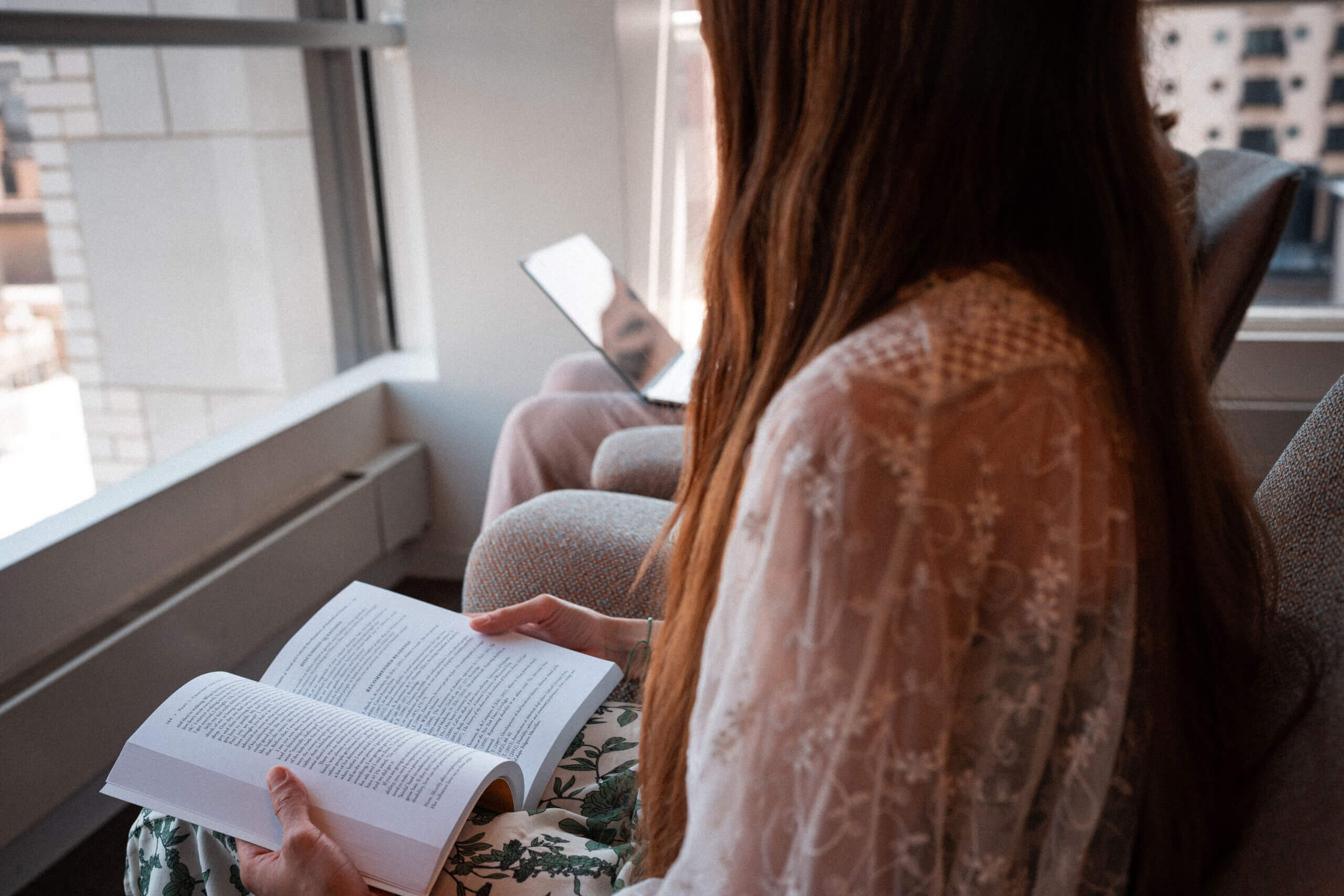 A woman with long brown hair reads a book while sitting on a chair by a window. Another person, partially visible, sits nearby using a laptop. City buildings can be seen outside the window.