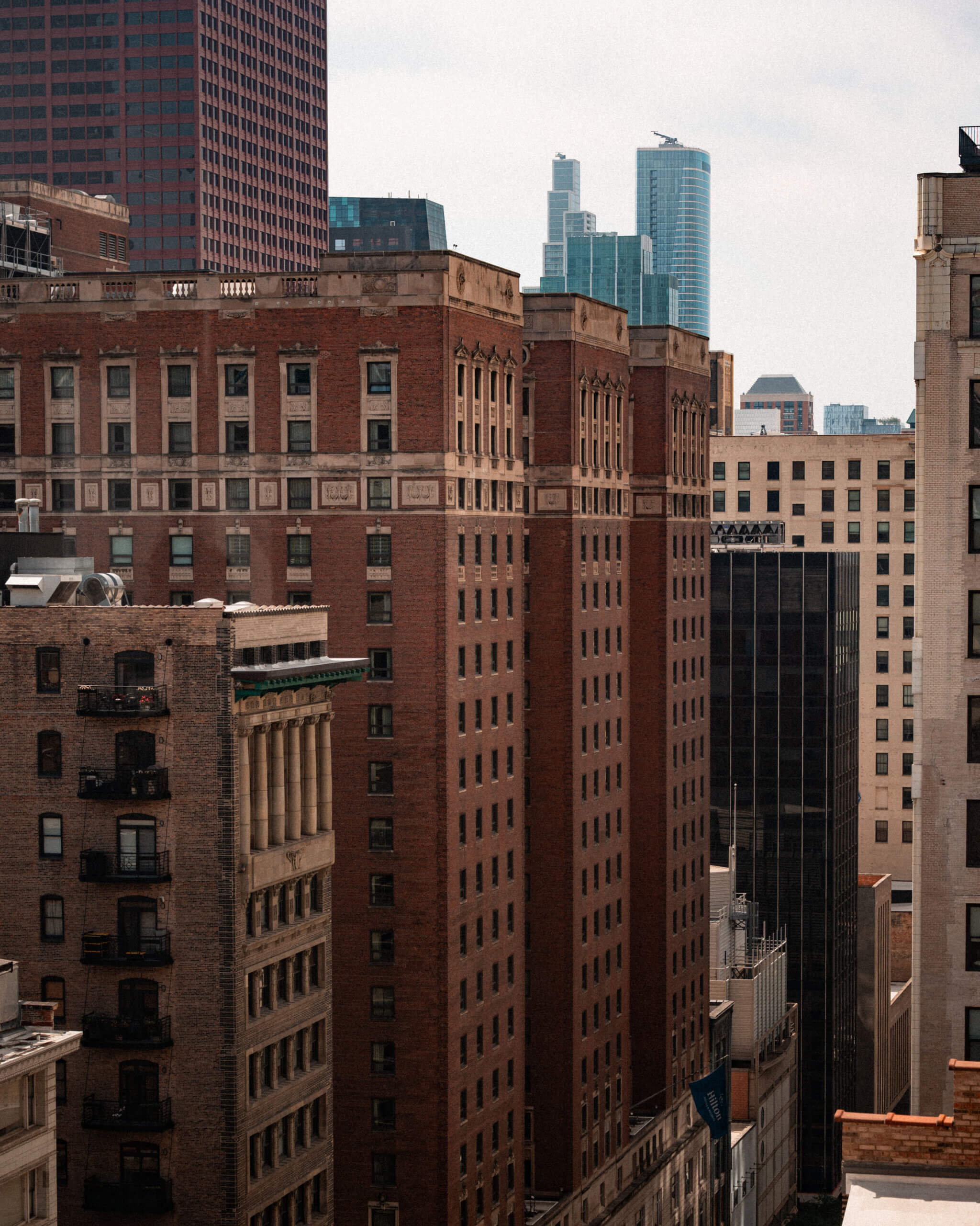 A cityscape showing tall brick and concrete buildings in the foreground with modern glass skyscrapers visible in the background under a cloudy sky.