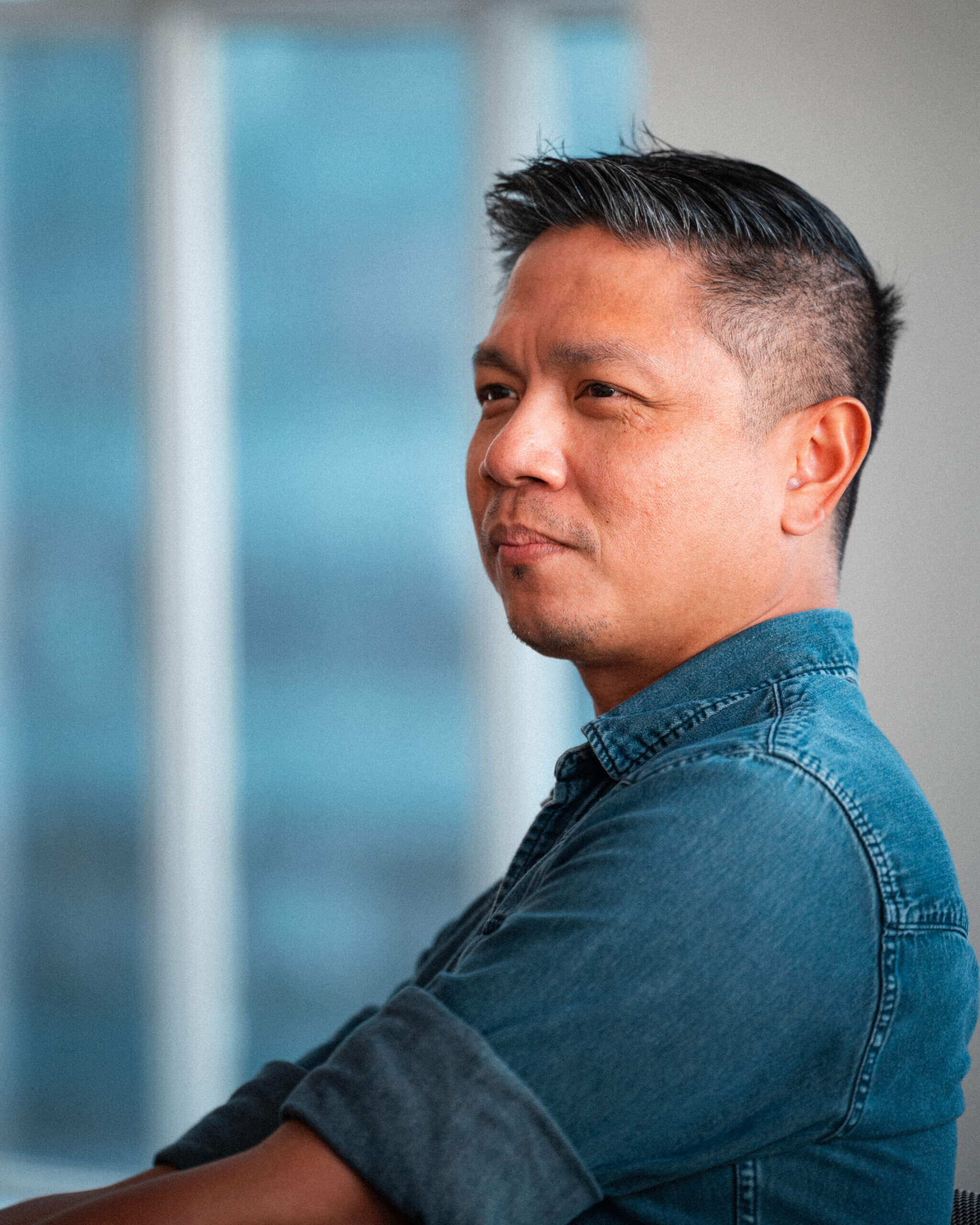 A man with short dark hair and a trimmed beard, wearing a blue denim shirt, is sitting in profile and looking thoughtfully into the distance. The background is softly blurred.