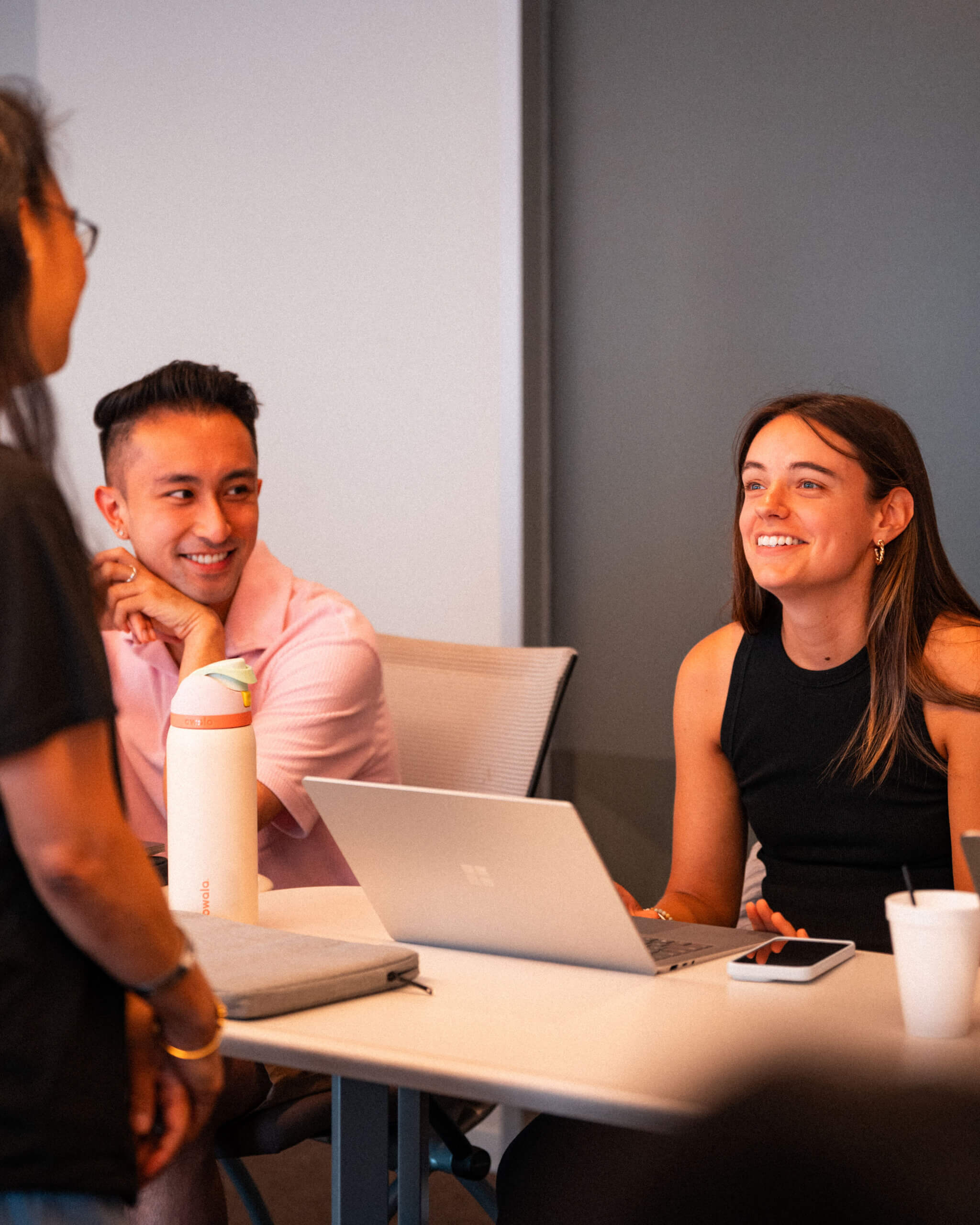 Two people sit at a table with a laptop, water bottle, and cup, smiling and talking to a standing person. The setting appears to be a casual office or meeting room.