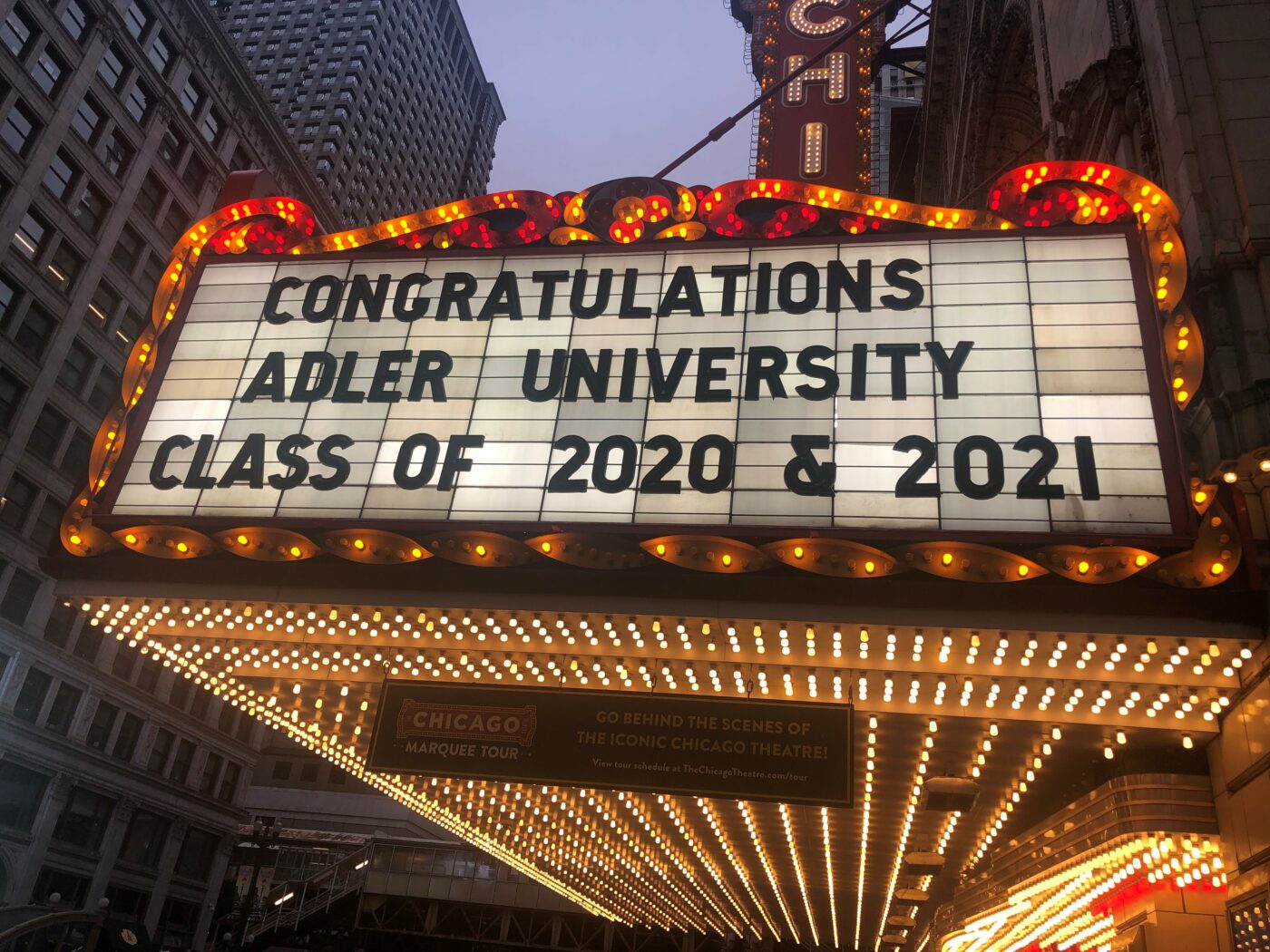A theater marquee reads, "Congratulations Adler University Class of 2020 & 2021," celebrating Chicago graduates, with lit bulbs and city buildings visible in the background.