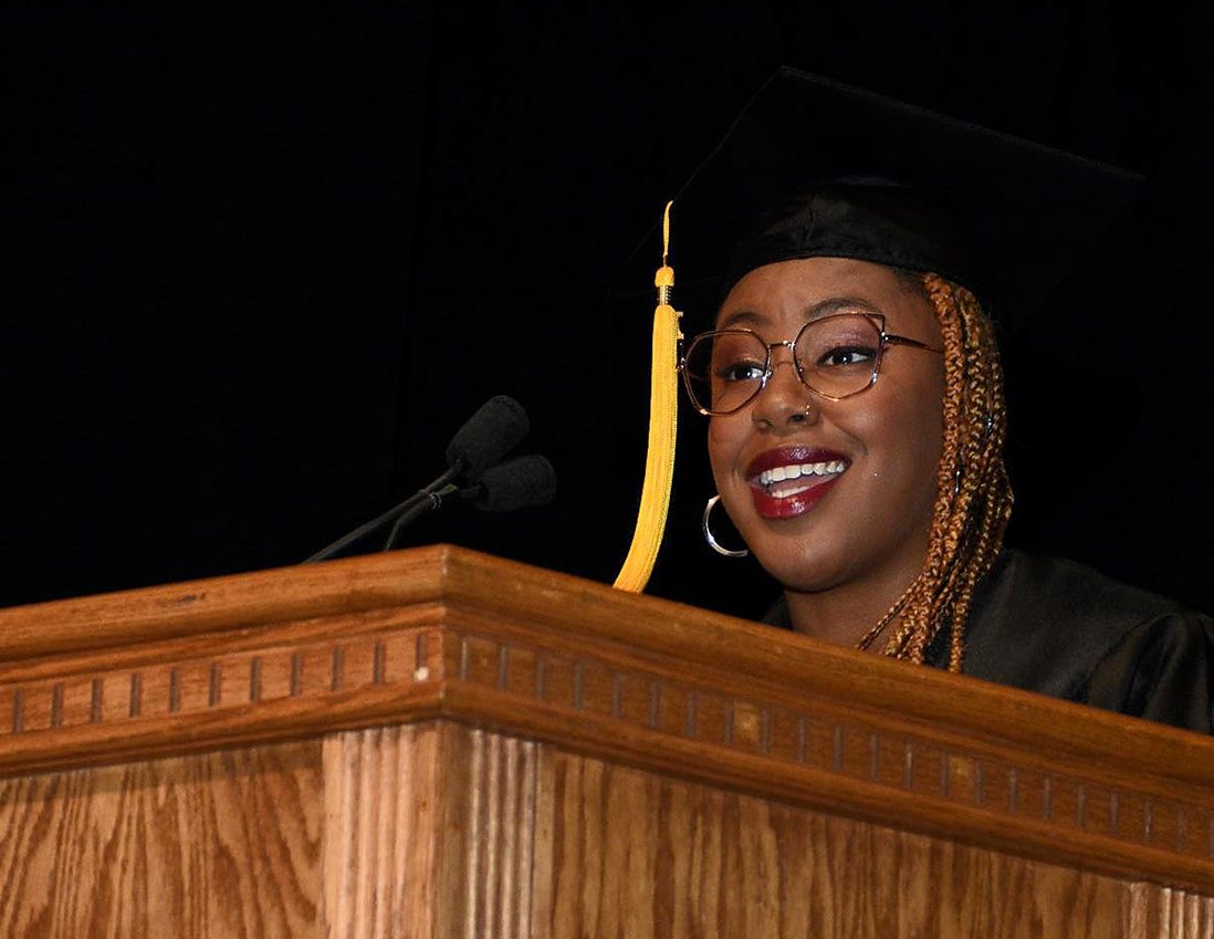 A diverse and inclusive Chicago graduate in graduation attire stands at a wooden podium, speaking into a microphone against a black background.