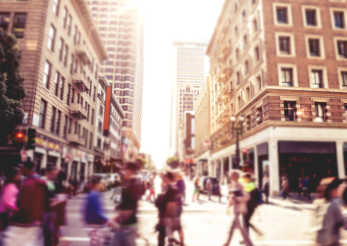 A busy city street with blurred pedestrians crossing at an intersection between tall buildings in daylight.