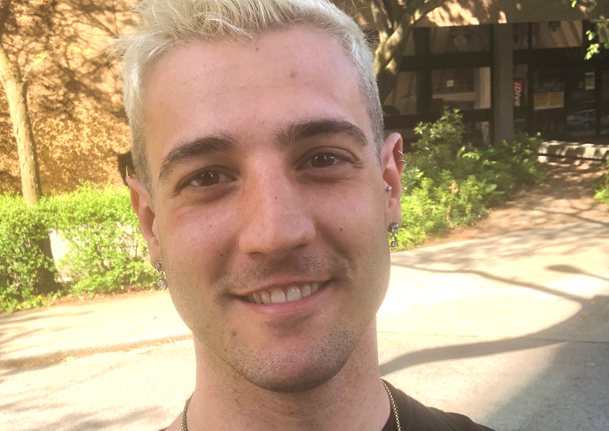 A person with short, bleached blonde hair and earrings smiles outdoors with trees and a building in the background, embodying resilience as vulnerable youth find ways to cope during the pandemic.
