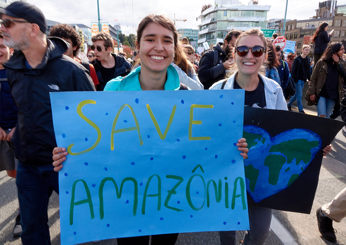 Two people at a protest embody climate activism, holding signs—one reading "SAVE AMAZÔNIA," another with a painted Earth inside a heart—spreading hope and inspiring action among a crowd on a city street.