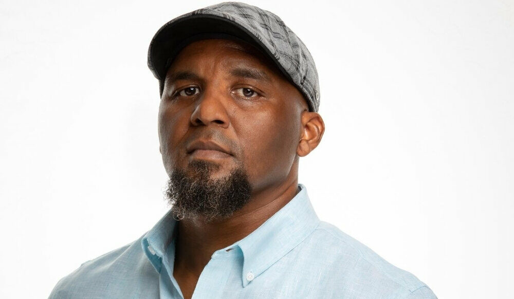 A man with a short beard and mustache, wearing a light blue shirt and a gray flat cap, stands in front of a plain white background.