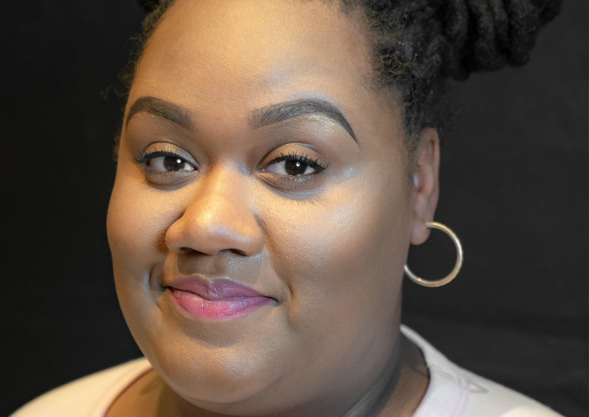 A woman with dark braided hair, hoop earrings, and a light pink top smiles softly against a dark background, embodying the strength of women shaping the next generation.