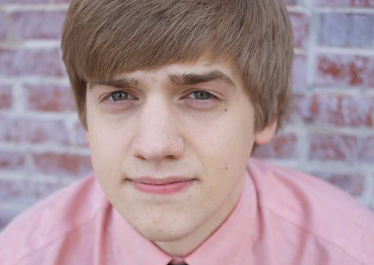 A young person with light brown hair wearing a light pink shirt stands in front of a brick wall, looking directly at the camera with a neutral expression, reflecting a calm and composed academic presence.