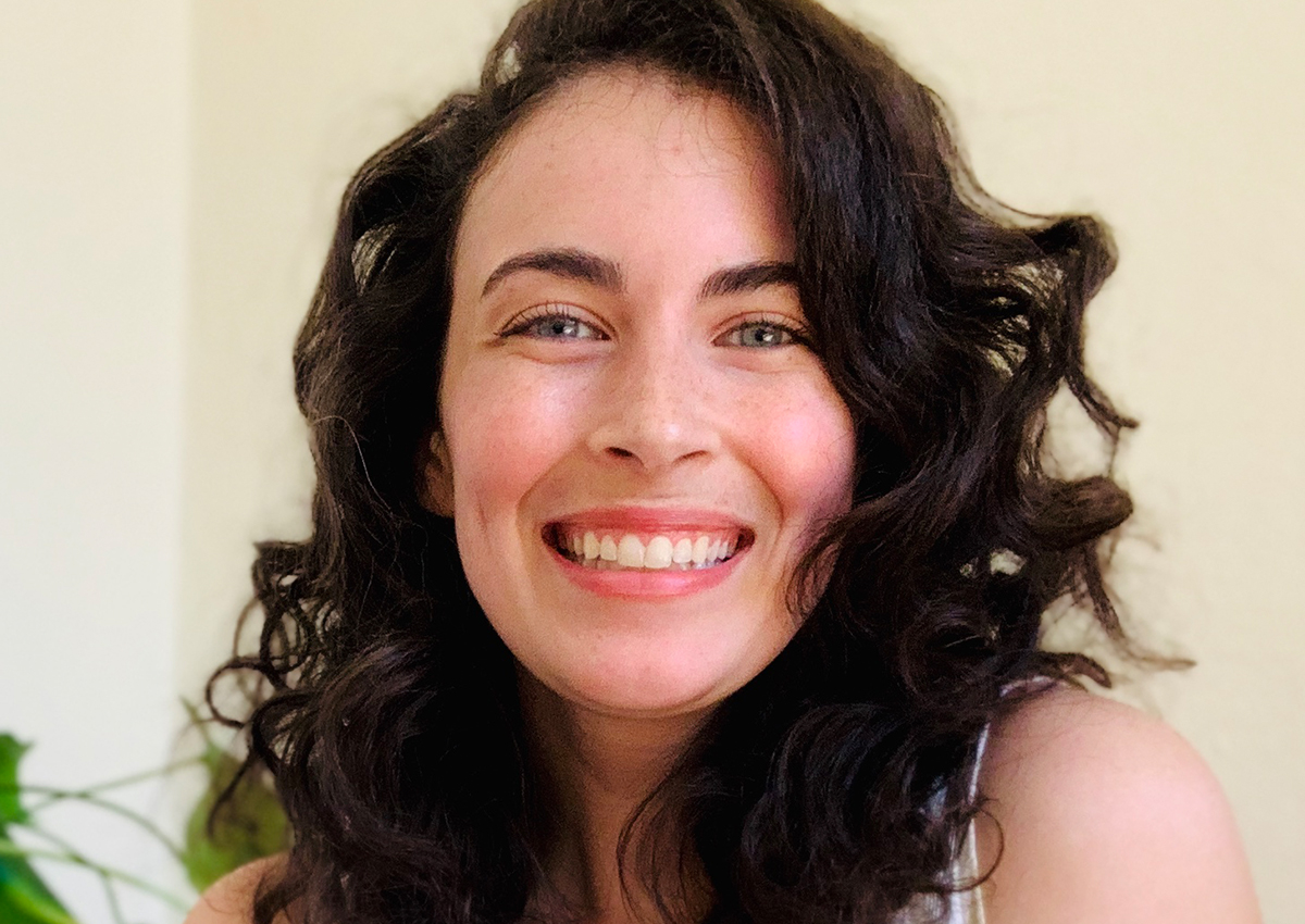 A person with curly brown hair and light skin smiles at the camera against a plain background, with a green plant partially visible, highlighting the importance of addressing rising risk factors related to domestic abuse.
