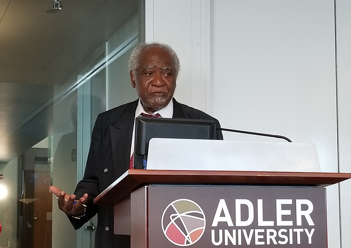 An older man in a suit speaks at a podium with a computer, in front of an Adler University Chicago Campus sign.
