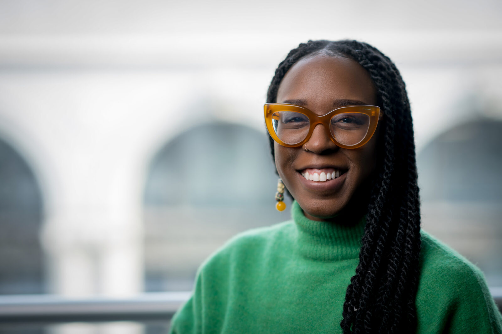 A person with long braids, wearing large brown glasses and a green sweater, smiles at the camera with a blurred background.