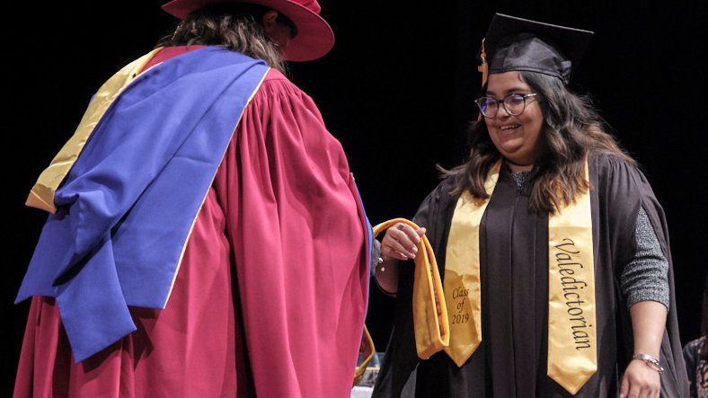 A Vancouver Graduate in cap and gown with a "Valedictorian" sash receives a cord from a faculty member in academic regalia, marking the privilege of achievement during the ceremony.