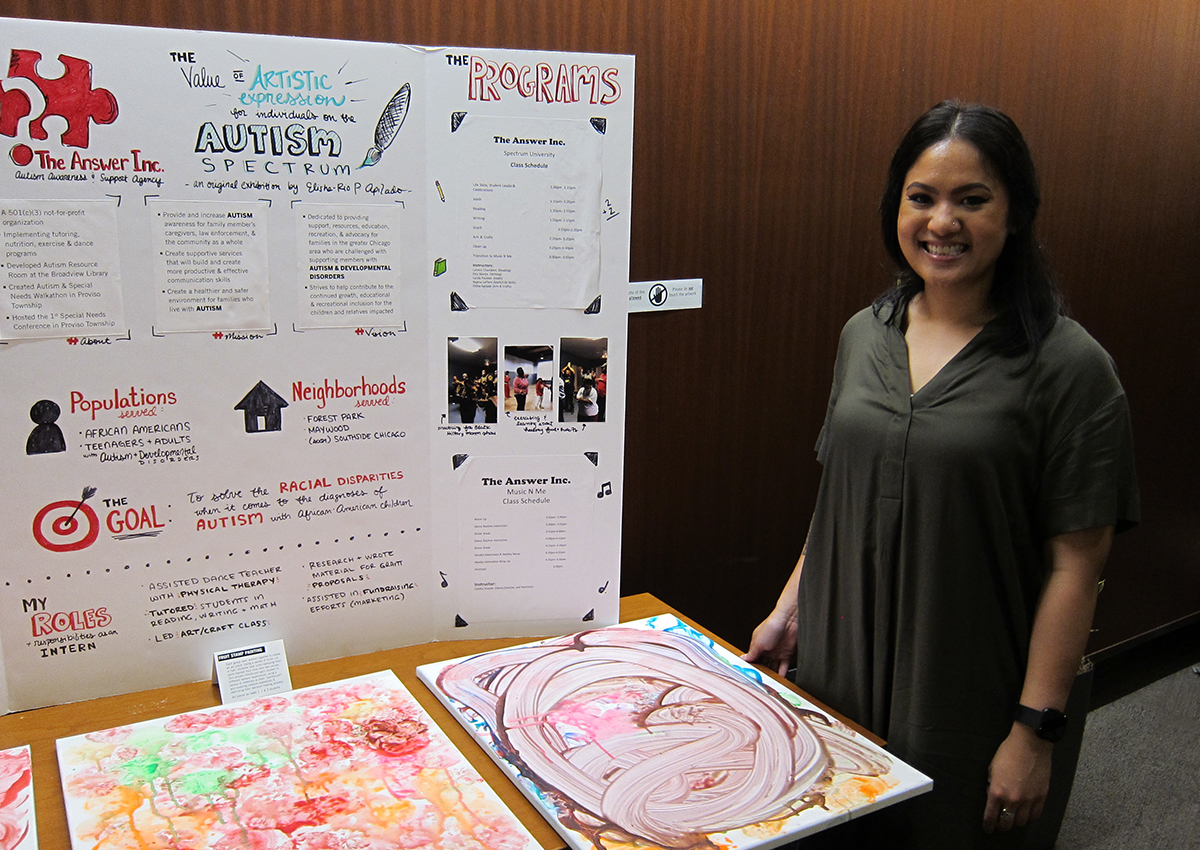 A woman stands next to a display about autism spectrum awareness and program information for teenagers and adults, with colorful artwork on the table in front of her.