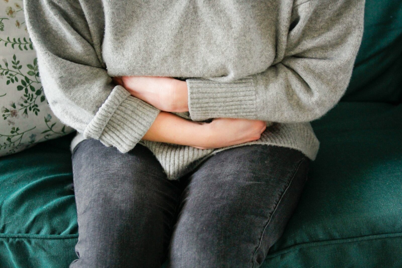 A person sits on a green couch, wearing a grey sweater and dark pants, holding their stomach as if experiencing pain or discomfort, possibly related to women’s reproductive health such as endometriosis.