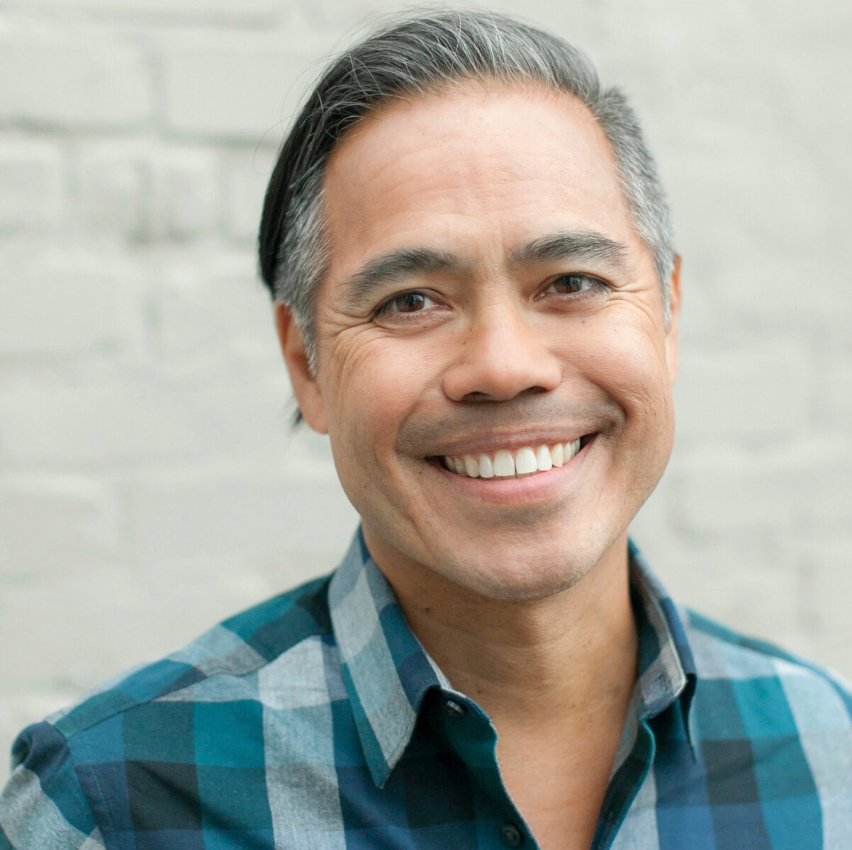 Dr. Evan Adams, a proud Vancouver Graduate known for his work in First Nation and Inuit Health, smiles at the camera in front of a light-colored brick wall, his short dark hair flecked with gray and wearing a blue plaid shirt.