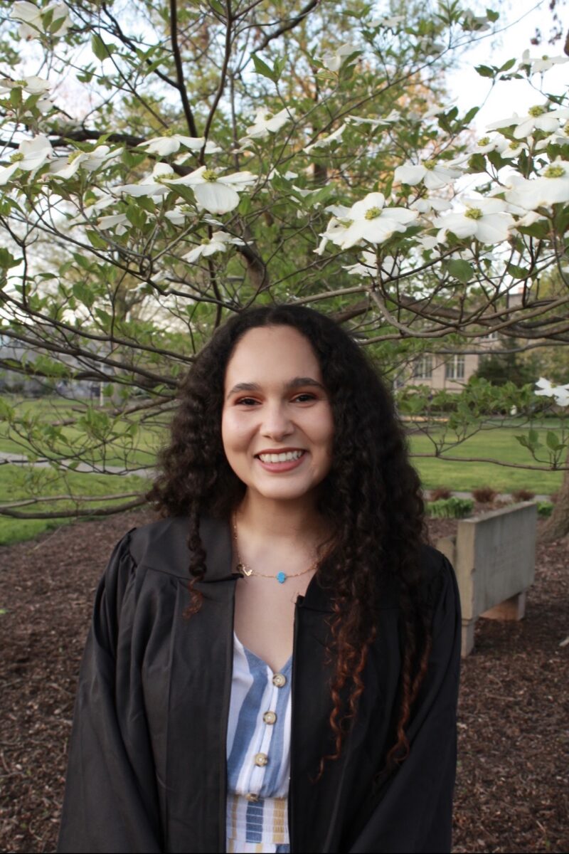A young woman with curly hair smiles warmly, wearing a graduation gown. She stands in front of a blossoming tree, conveying happiness and achievement.