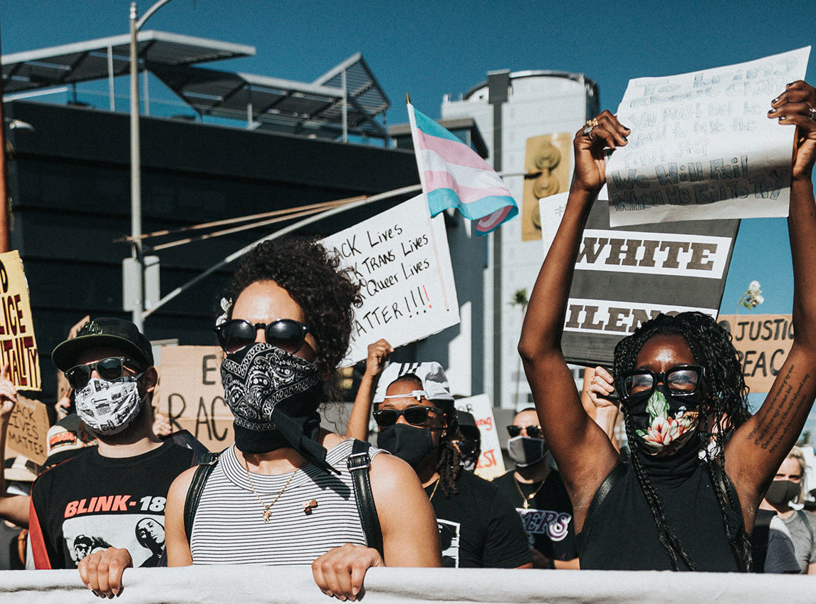 A group of masked protesters hold signs at a demonstration, with a transgender pride flag visible in the background, highlighting solidarity in coping with stress and standing against racial injustice.
