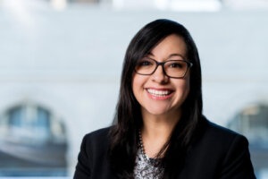 Flor Esquivel sits in front of a large window with buildings blurred in the background. She is wearing a blouse and blazer.