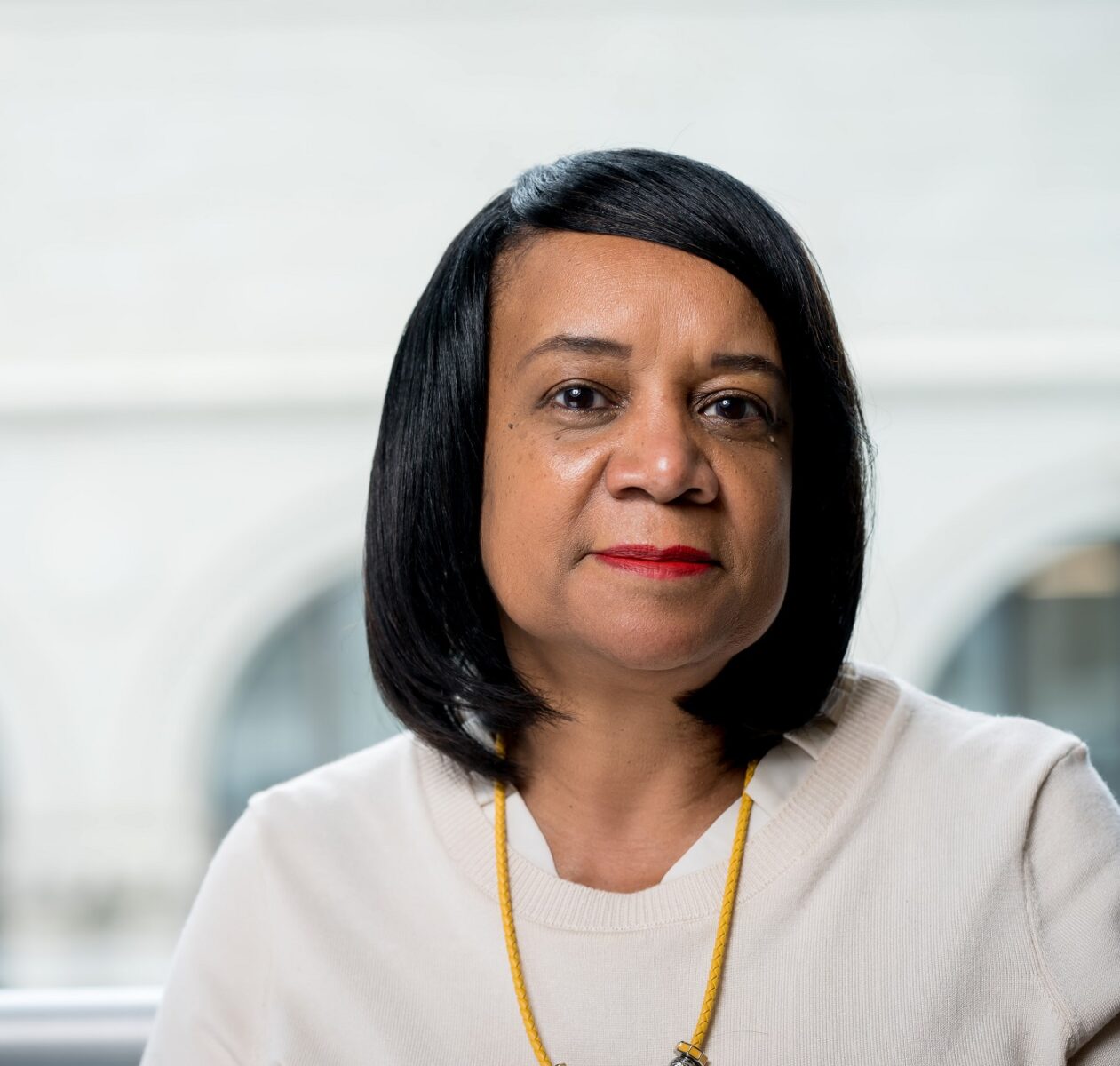 A woman with straight black hair and red lipstick, wearing a cream sweater and yellow necklace, sits in front of a blurred light-colored background, appearing ready to discuss community psychology or review an eBook.