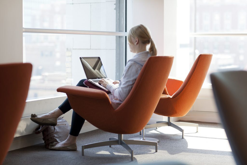 A person sits in an orange chair by a large window, using a laptop on their lap in a brightly lit modern indoor space, working on a digitization project for archival videos.