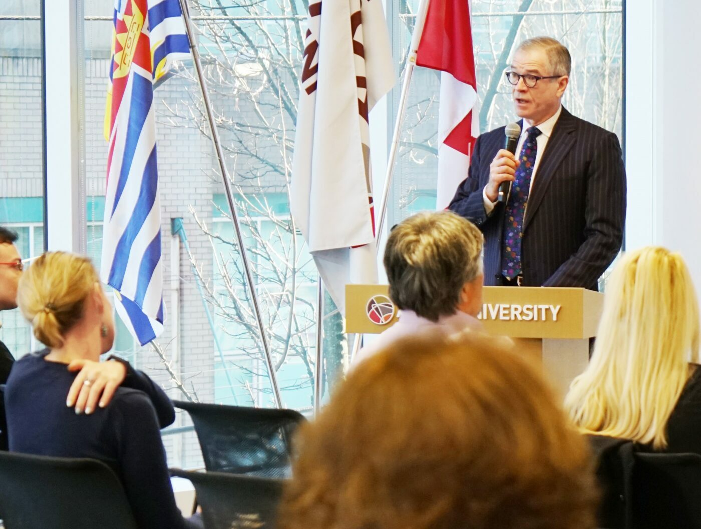 Bradley O’Hara speaks at a podium labeled "UNIVERSITY" to an attentive audience, with flags in the background, representing Adler Vancouver and addressing the Degree Quality Assessment Board.