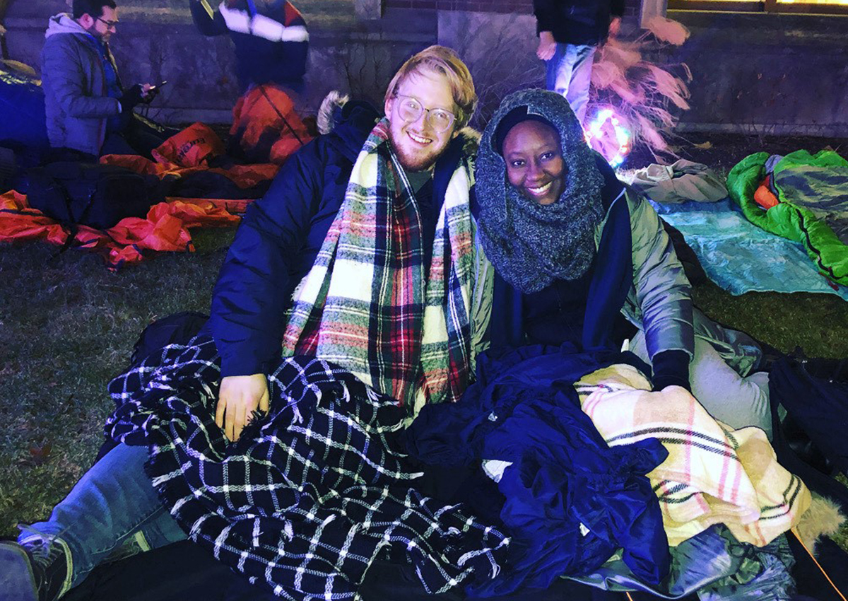 Two people sit on the ground outdoors at night, bundled in scarves and blankets, smiling at the camera with others and sleeping bags in the background—raising awareness about different forms of homelessness.