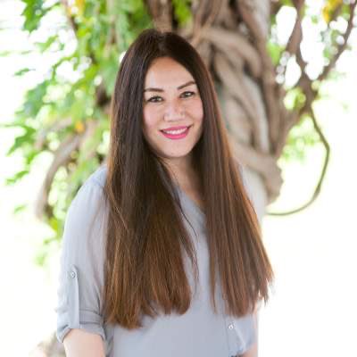A woman with long brown hair, wearing a light gray blouse, stands outdoors in front of a tree with twisted branches and green leaves at Adler University, recipient of the 2021 APA Presidential Citation.