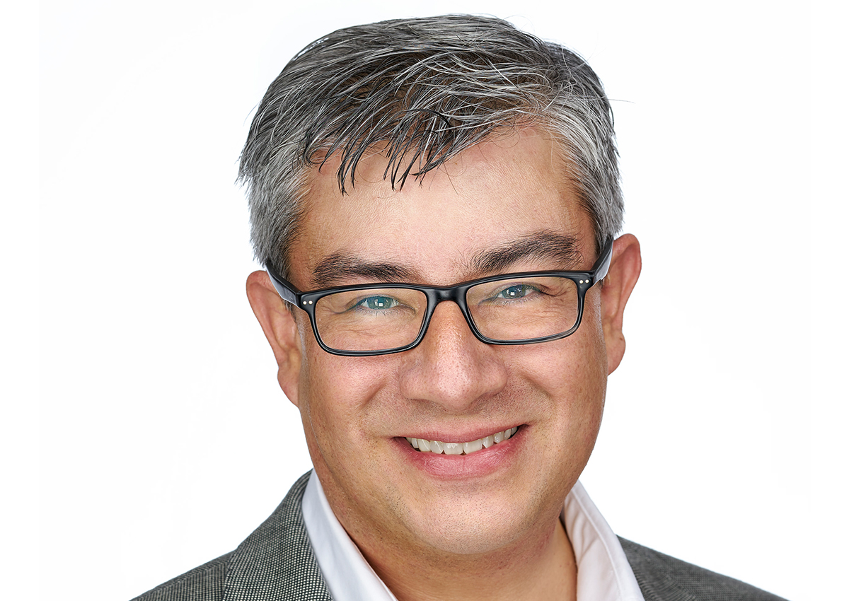 A middle-aged man with gray hair, wearing glasses, a gray blazer, and a white shirt, smiles at the camera against a plain white background, embodying the spirit of workers rights and advocacy.