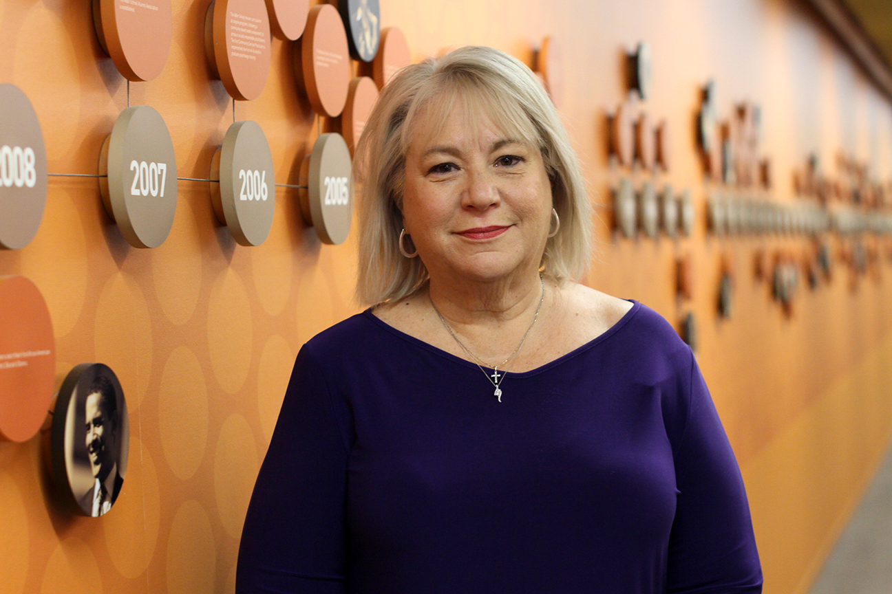 A woman with shoulder-length gray hair wearing a purple top stands in front of an orange wall decorated with a timeline and circular plaques highlighting community service.