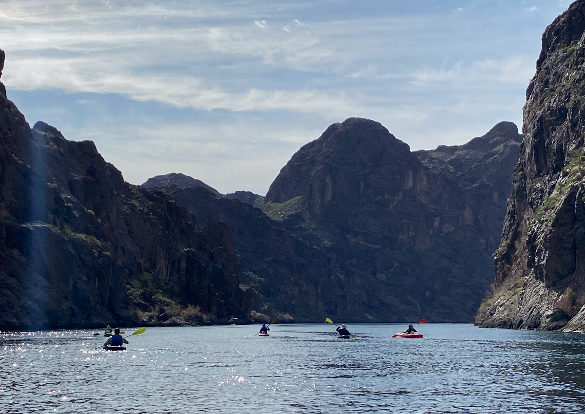 Four people kayak through a wide river canyon surrounded by steep rocky cliffs under a partly cloudy sky, embracing the power of nature.