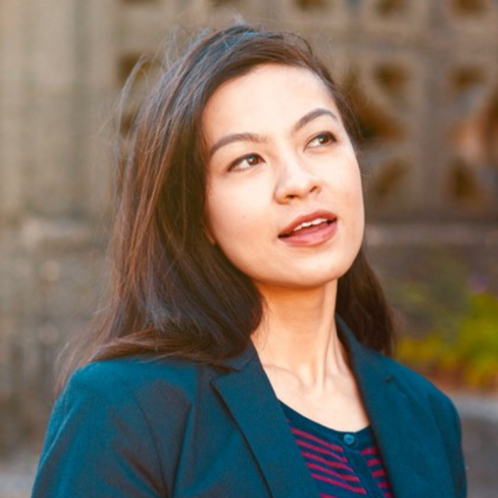 A woman with long dark hair, wearing a dark blazer over a striped shirt, looks slightly upward with a thoughtful expression, as if reflecting on ethical fashion practices. The background is blurred.