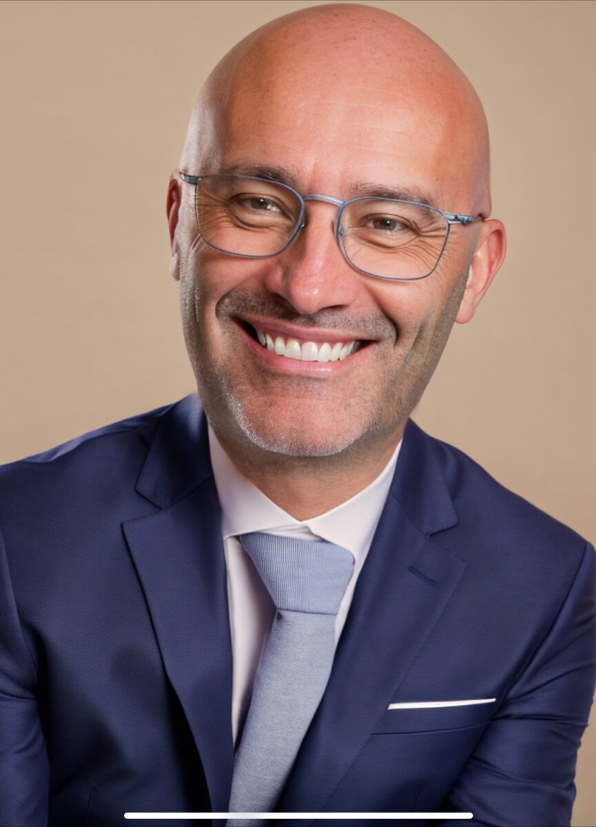 A bald man wearing glasses and a blue suit with a white shirt and light blue tie, smiling at the camera against a beige background—Jason Walker, contributing writer for Forbes.